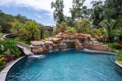 Waterslide framed by vibrant tropical plants and stonework in a custom outdoor pool setting.