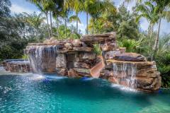 Custom luxury waterslide built into a natural rock pool with tropical landscaping and cascading waterfalls.